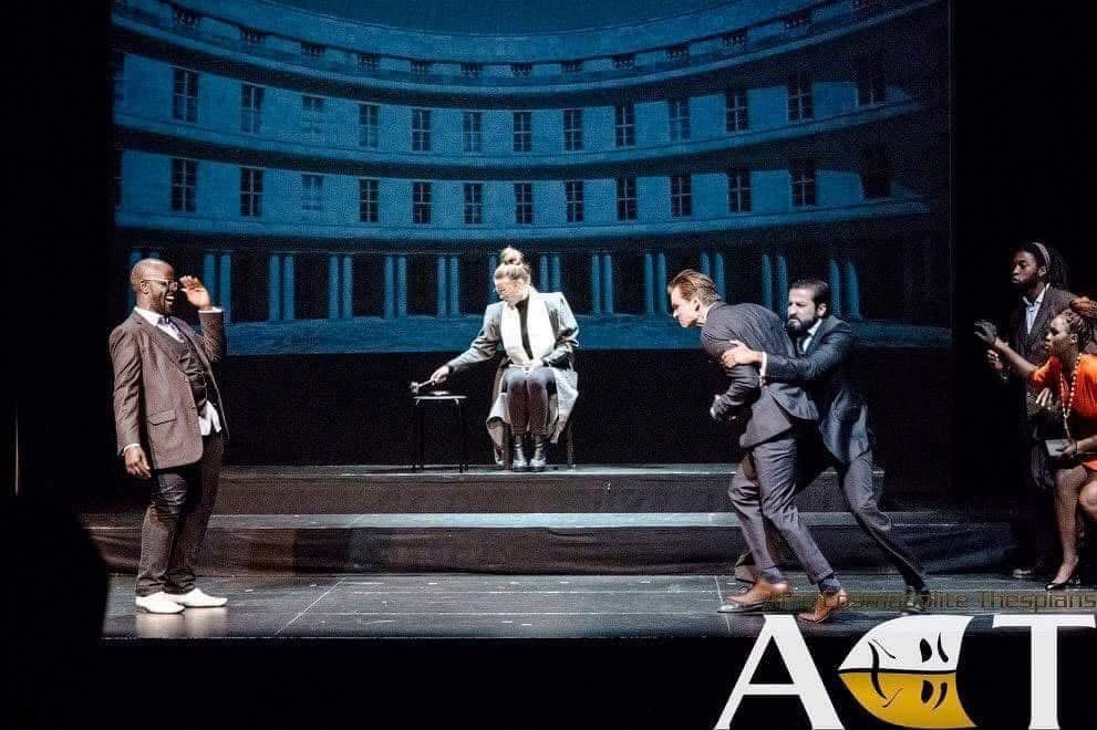 Wide courtroom tableau from The Merchant of Venice with Shylock facing the court as others surge around Antonio.