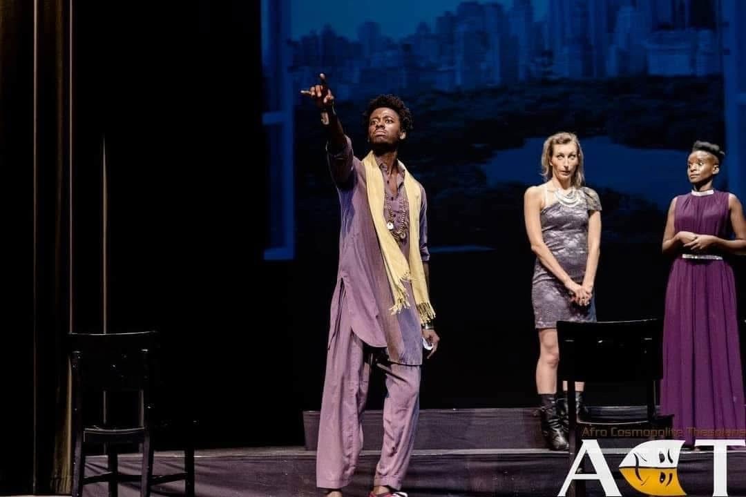 Production still from The Merchant of Venice showing a central figure pointing upward while two women stand behind him on stage.