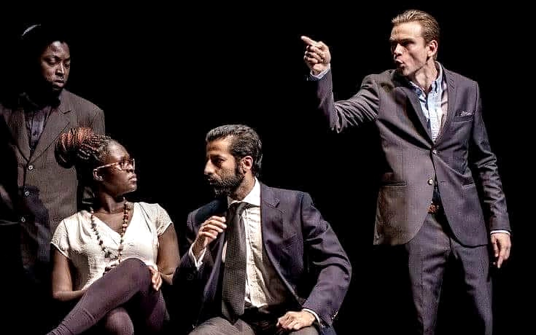 Production still from The Merchant of Venice showing a central figure pointing across the stage while two women stand behind.