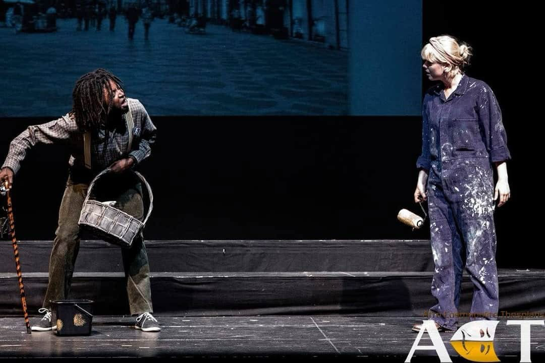 Launcelot in a comic stage still from The Merchant of Venice holding a bucket and stick while arguing with another character.