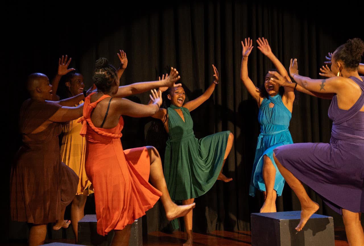 Women in colored dresses rising together in a celebratory circle.